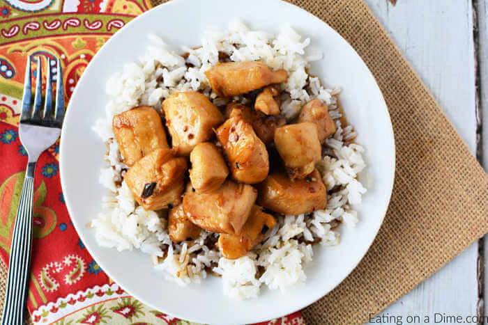 Close up image of a white plate of teriyaki chicken on a bed of rice. It is on a place mat with a fork. 