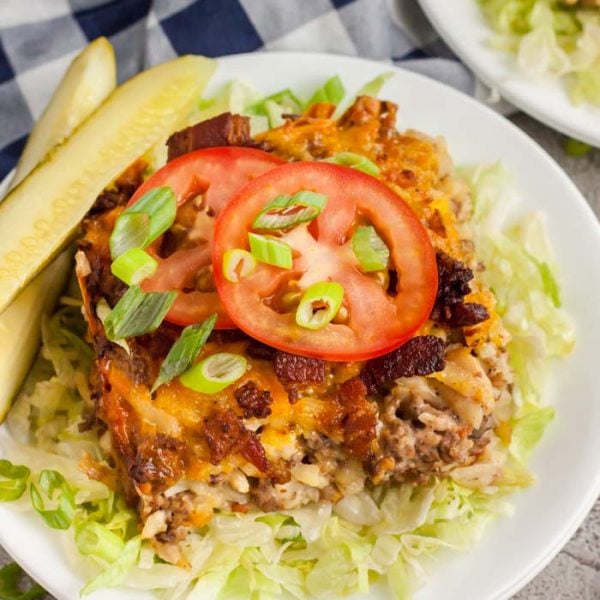 A serving of bacon cheeseburger casserole topped with sliced tomatoes, green onions, and melted cheese, served on a bed of shredded lettuce with crispy bacon and two pickle spears on the side.