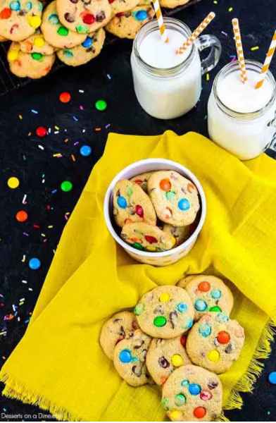 Photo of yellow napkin with M&M Cookies on it surrounded by cups of milk and more cookies.