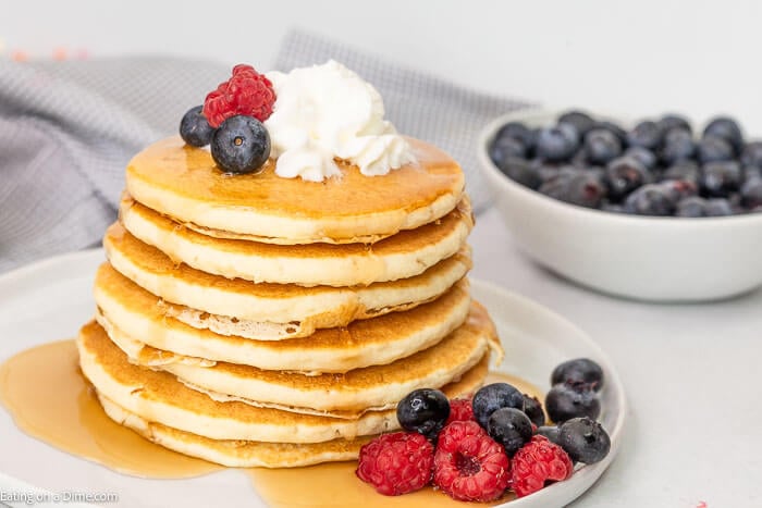 A white plate full of cooked pancakes stacked on top of each other topped with syrup, whipped cream and fresh berries 