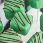 Close-up of Minecraft Cake Pops: square cake pops covered in green icing and drizzled with chocolate, displayed on white sticks.