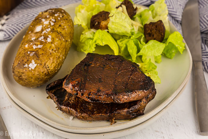 2 steaks on a cream color plate with a baked potato and salad next to them. 