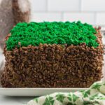 A square Minecraft Birthday Cake covered in chocolate crispy cereal on the sides, with green frosting piped on top to resemble grass, sits on a white plate with a green and white towel nearby.