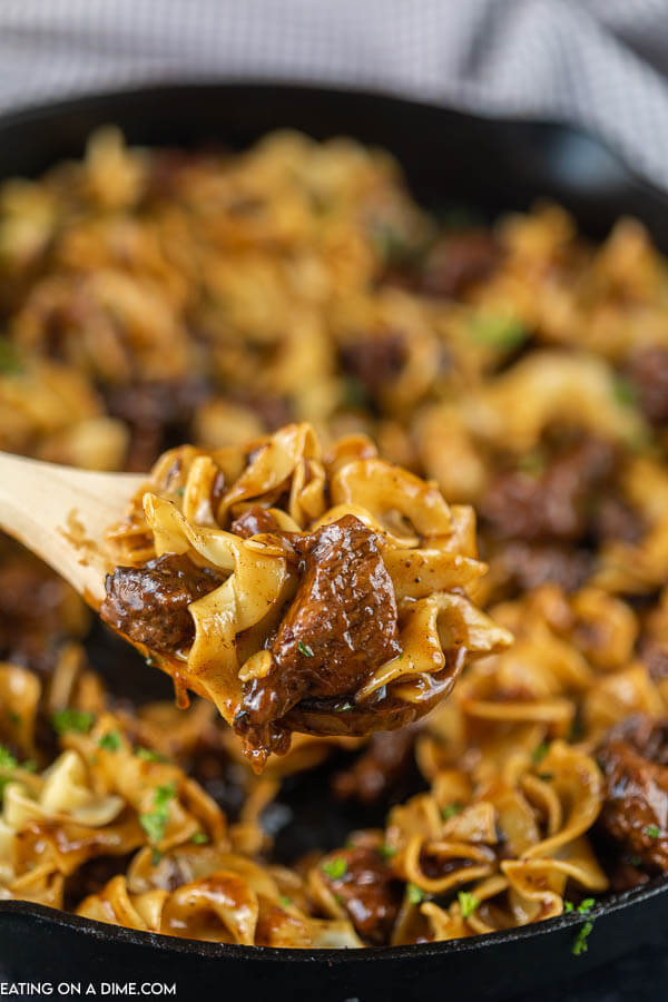 Close up image of beef and noodles in a iron skillet with a serving on a wooden spoon. 