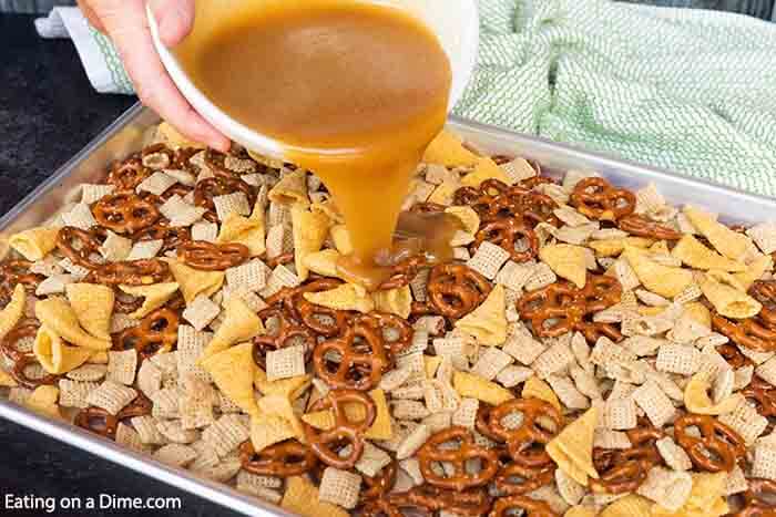 Pouring the brown sugar mixture over the cereal, pretzels, and bugels on a baking sheet