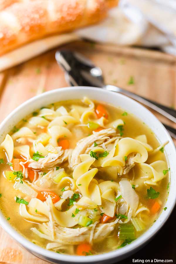 A bowl of chicken noodle soup with bread and spoons above it. 