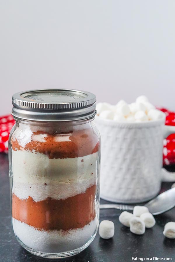 Close up image of hot chocolate mix layered in a mason jar with a mug of hot chocolate with marshmallows. 