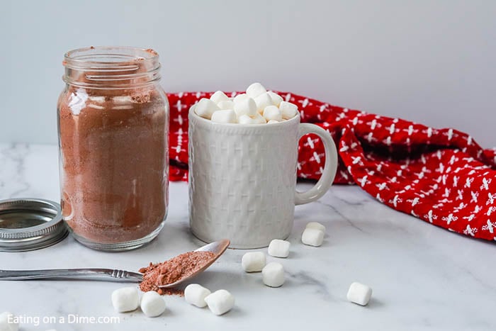 Close up image of hot chocolate mix in a mason jar with a spoon. Also a white mug with hot chocolate with marshmallows. 