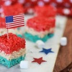 Red, white, and blue Rice Krispie Treats shaped like hearts, topped with a small American flag, displayed on a marble surface with star decorations. The treats are festive for 4th of July celebrations.