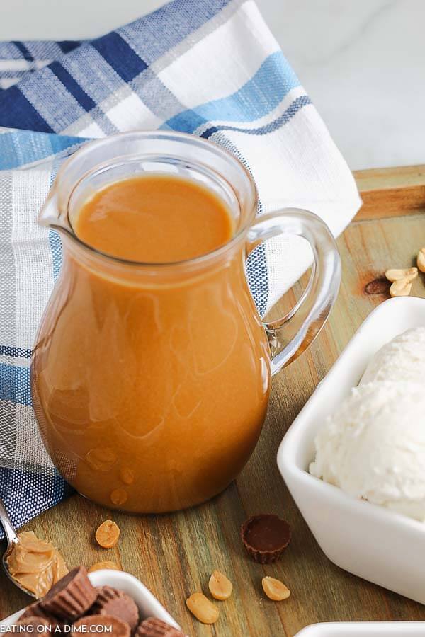 A small glass pitcher of the peanut butter sauce in it next to vanilla ice cream on a wooden platter. 
