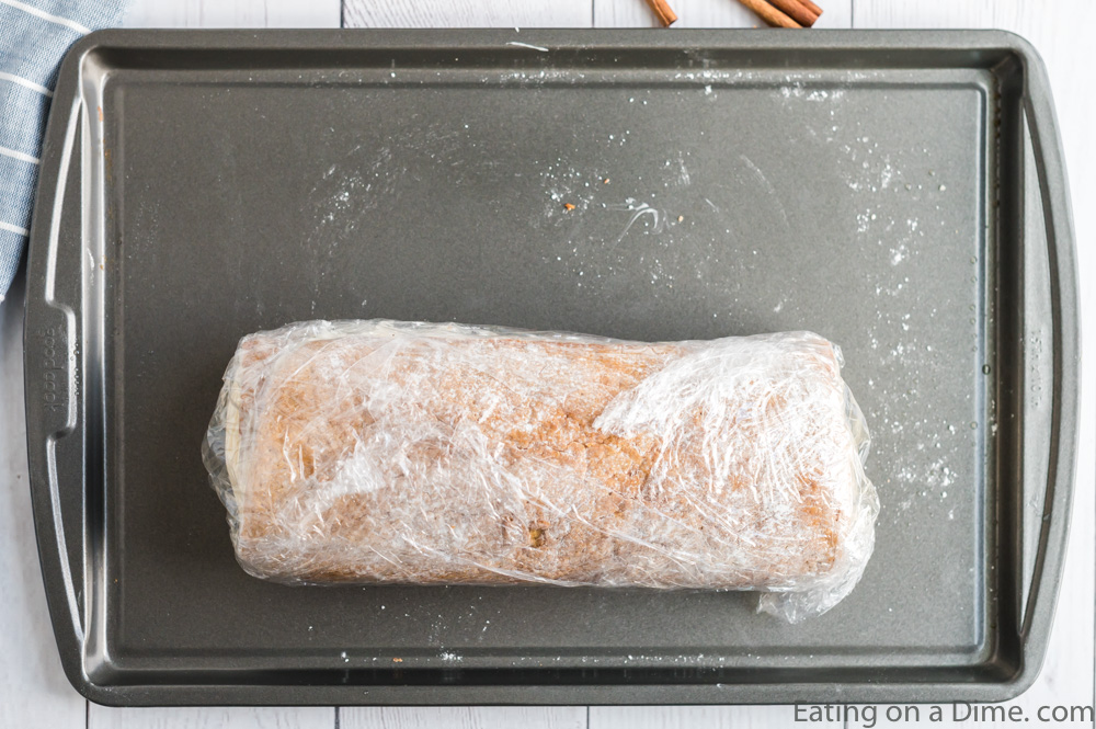 Close up image of rolled pumpkin roll wrapped in saran wrap on a cookie sheet. 