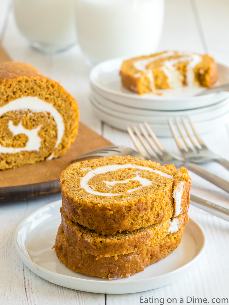 Close up image of pumpkin roll stacked on a white plate with slices in the background on a cutting board. 
