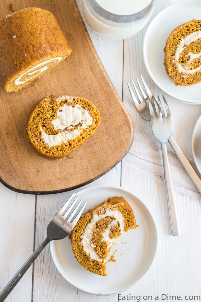 Close up image of a slice of pumpkin roll on a white plate in a slice on a cutting board with two silver forks.
