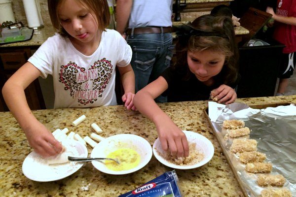 two girls dipping the cheese sticks in bread crumbs and eggs and placing on the baking sheet
