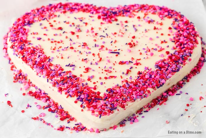 A heart shaped cake decorated with pink, red and purple sprinkles. 