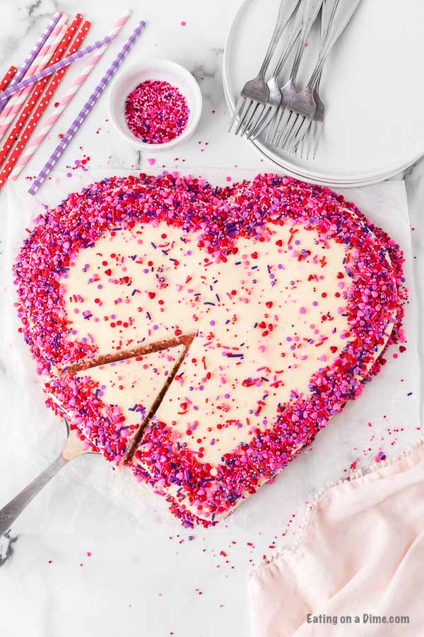 A heart shaped cake decorated with pink, red and purple sprinkles with 1 piece cut out of it and ready to be served. 