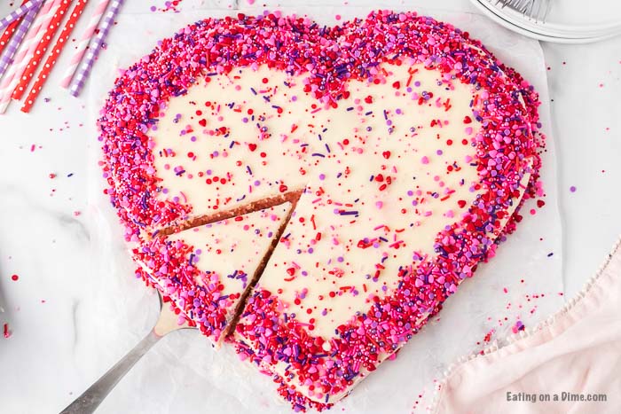 A heart shaped cake decorated with pink, red and purple sprinkles with a piece being cut from the cake 