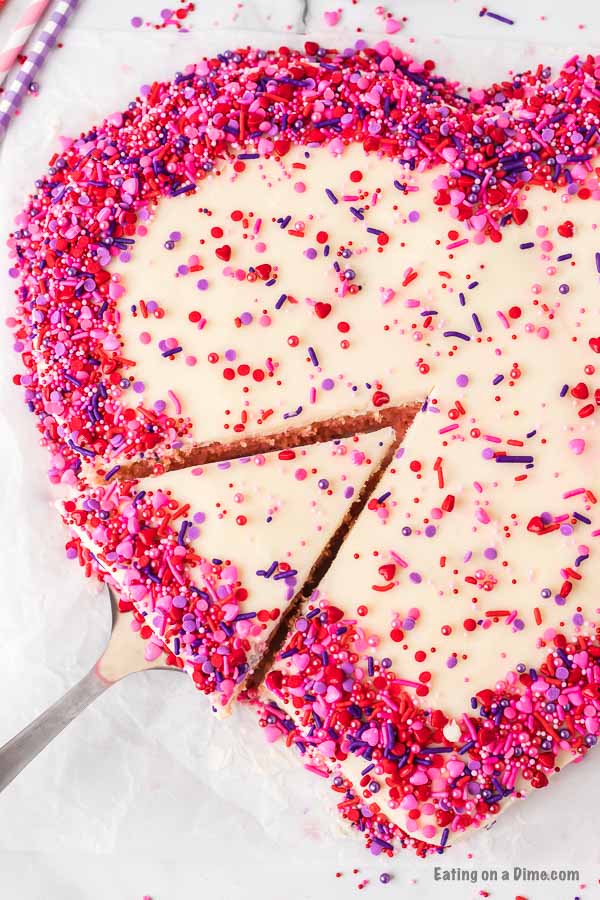 Close up of a heart shaped cake decorated with pink, red and purple sprinkles with a piece being removed from the cake. 