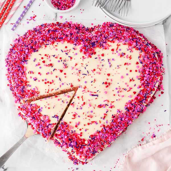 A heart shaped cake decorated with pink, red and purple sprinkles. 