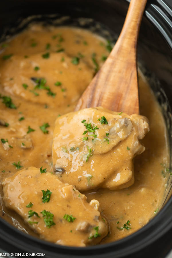 Close up image of smothered pork chops in a slow cooker with a wooden spoon. 