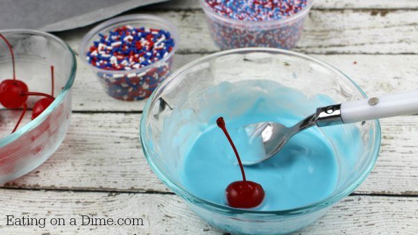 The light blue candy melts melted in a bowl and the cherry is being dipped in the candy. 