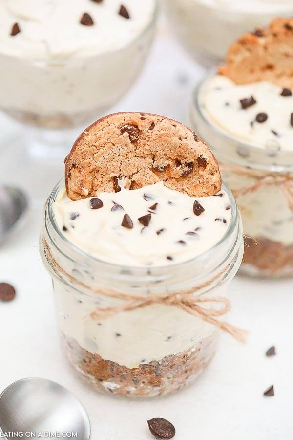 A jar with the no bake chocolate chip cookie cheesecake in it topped with a cookie. There are other jars in the background as well. 