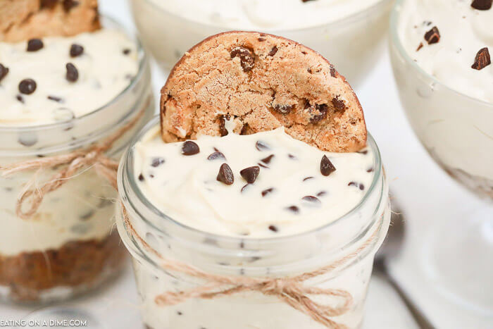 A jar with the no bake chocolate chip cookie cheesecake in it topped with a cookie with twine tied around the jar. 