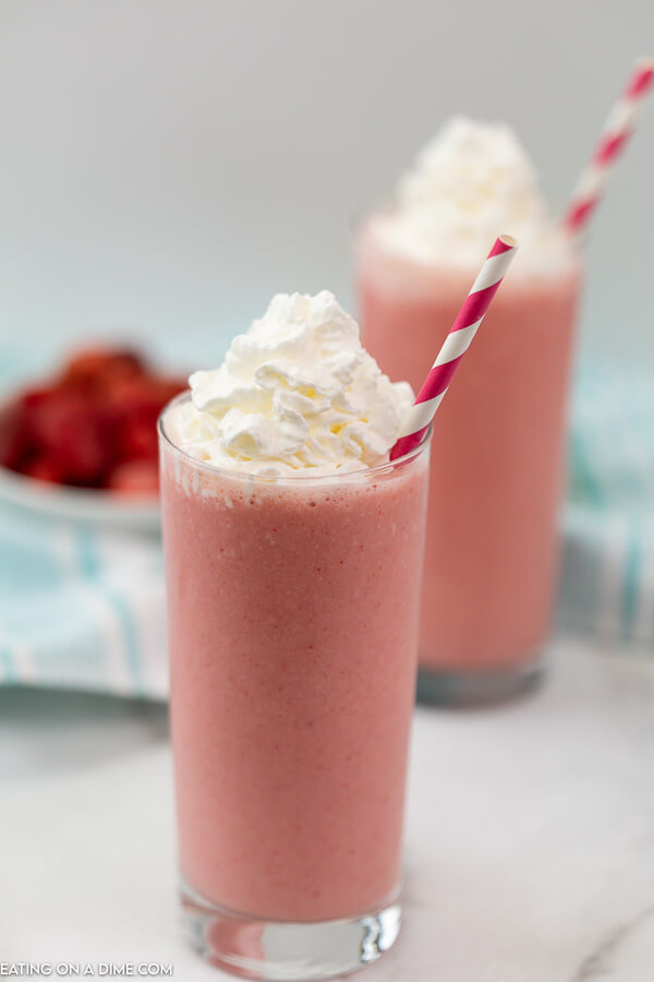 Strawberry Smoothie with whipped cream on top and a straw in it.  Then a smoothie in the background and strawberries in the back ground as well. 