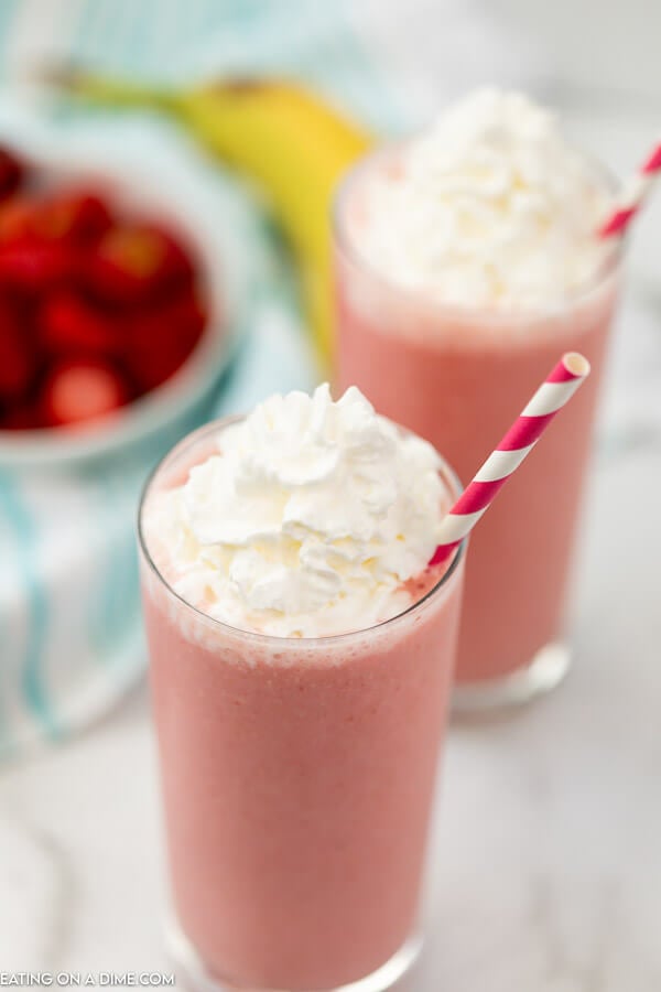 The top view of a Strawberry Banana Smoothie topped with whipped cream with a straw in it with a smoothie in the back ground with strawberries in the back ground too 