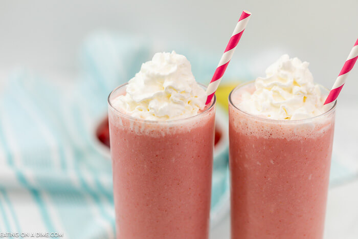 2 Strawberry Smoothies with whipped cream and straws in it with strawberries and lemons in the back ground and a blue linen. 