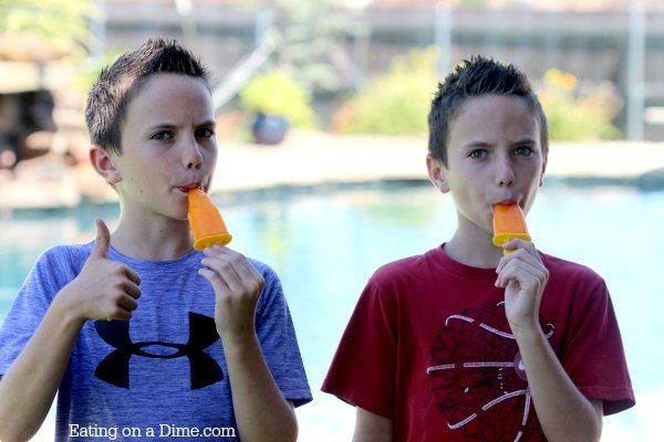 2 boys standing in front of a pool eating jello popsicles. 