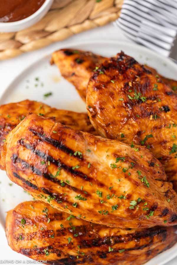 5 pieces of grilled chicken on a white plate and there is a small bowl of barbecue sauce in the background as well. 