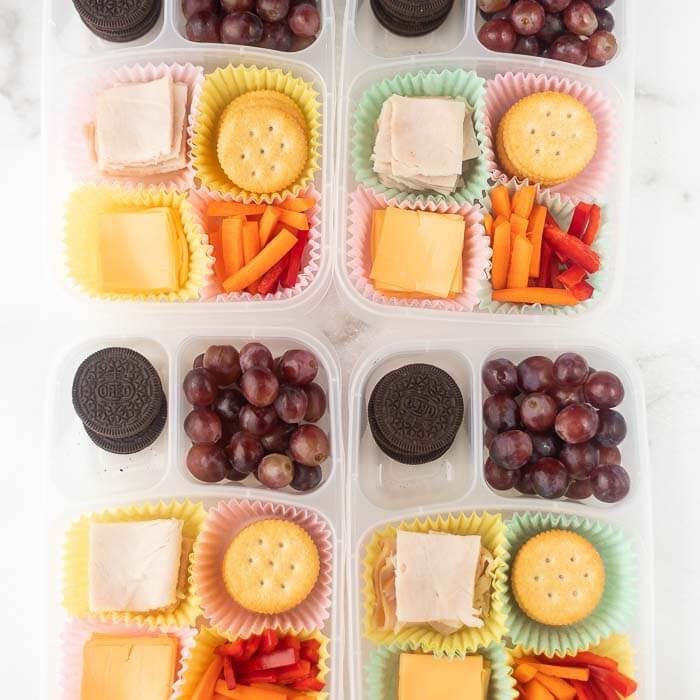Close up of containers with meat, cheese, carrots, crackers, oreo, and grapes. 