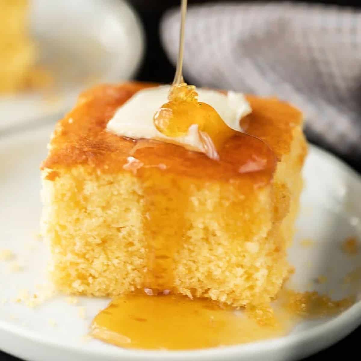 A square piece of cornbread topped with a pat of butter, with honey being drizzled over it, served on a white plate—perfect for anyone seeking the ultimate Honey Cornbread Recipe.