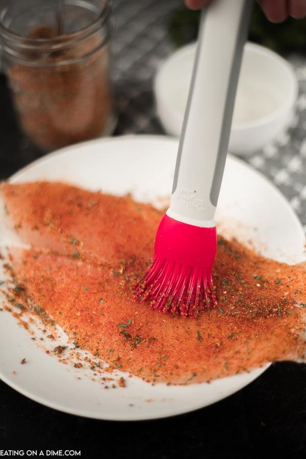 A red basting brush rubbing Blackened seasoning on a piece of tilapia on a white plate with blackened seasoning behind it 