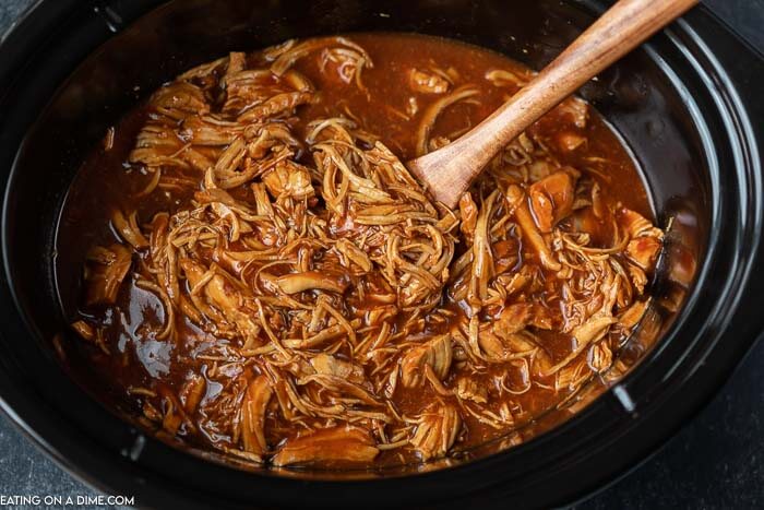 BBQ shredded chicken in a black crock pot with a wooden spoon scooping some of the chicken out of the crock pot. 