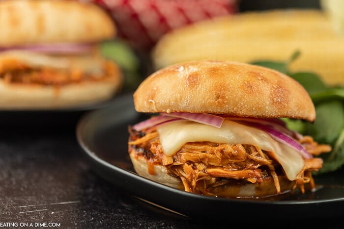 A BBQ pulled chicken sandwich topped with melted Monterey Jack cheese and sliced red onions on a black plate with salad next to it on the plate. 