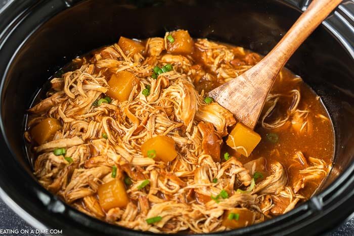 Hawaiian BBQ Chicken in a Black Crock Pot with a wooden Spoon in the chicken mixture 