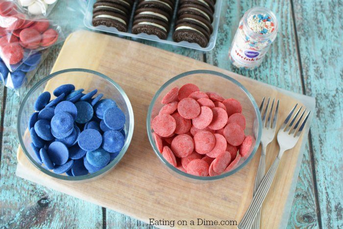 A bowl of blue and red chocolate melts on a cutting board lined with parchment paper. Two forks and a package of oreos
