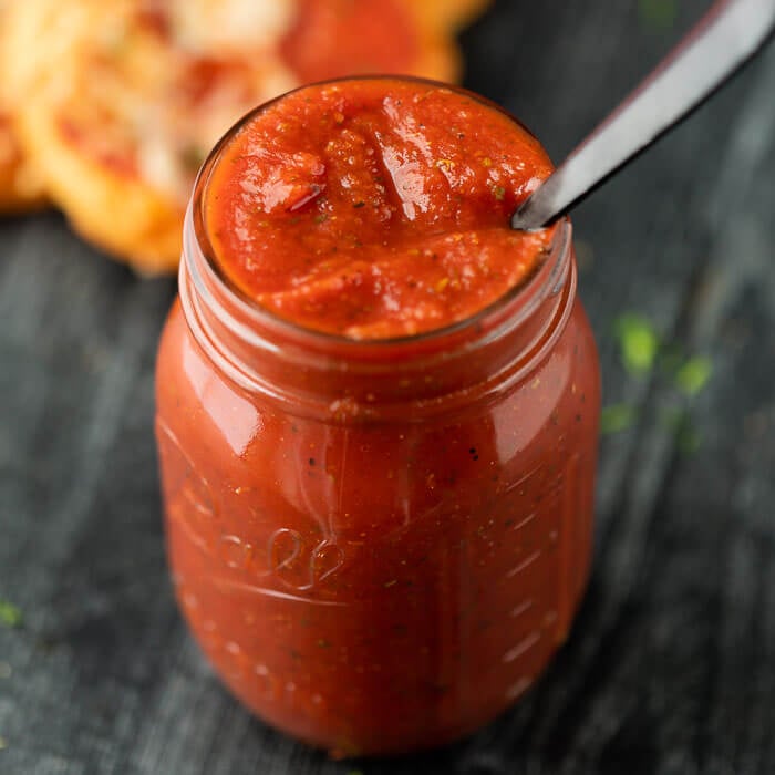 Pizza sauce in a mason jar with a silver spoon in it. with a pizza in the back ground 