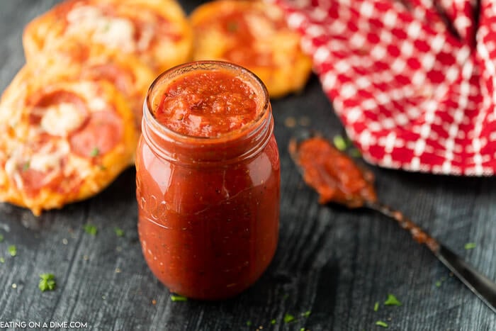 Pizza sauce in a mason jar with a messy spoon next to it and pizzas and red linen in the back ground 