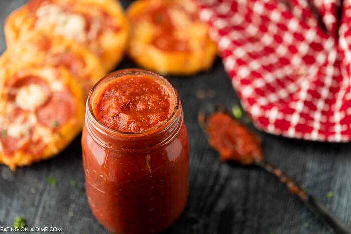 Pizza sauce in a mason jar with a messy spoon next to it with multiple mini pizzas in the back ground and a red and white linen in the back ground as well. 
