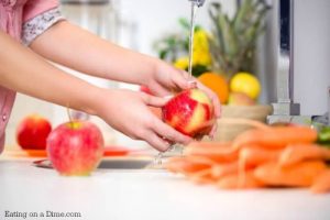 An overview of a countertop with lots of fruit and veggies on the countertop with a person washing an apple in a sink faucet 