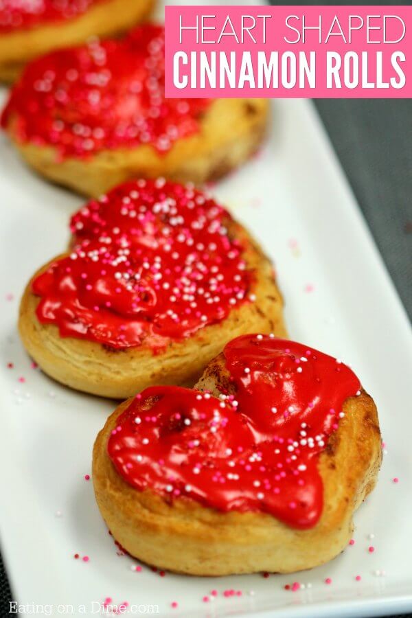 Heart shaped cinnamon rolls topped with red icing and Valentine's Day sprinkles on a white platter 