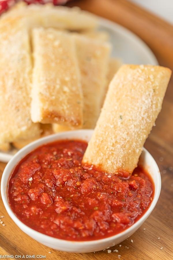 A Little Caesar's Crazy Bread Stick being dipped into a bowl of pizza sauce with the plate full of crazy bread behind it.  