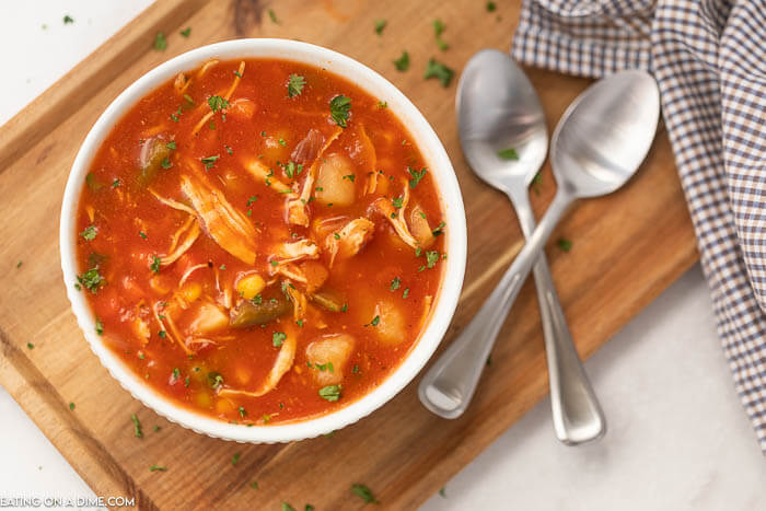 A bowl of chicken vegetable soup in a white bowl on a wooden platter with 2 silver spoons next to it. 