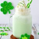A light green milkshake in a mason jar, topped with whipped cream and green sprinkles, with two green-striped straws. Shamrock decorations hint at a festive St. Patrick's Day and the classic McDonald's Shamrock Shake recipe inspiration.