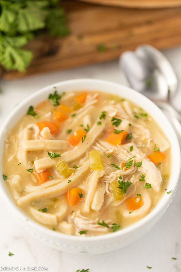 Close up image of chicken noodle soup in a white bowl on a table with two silver spoons. 