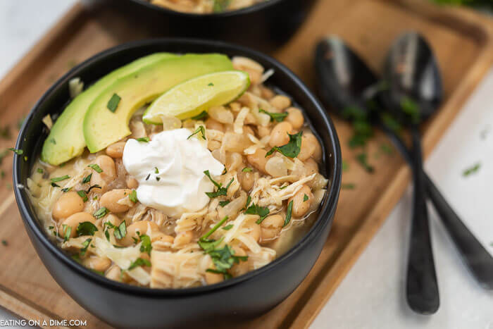 Close up image of a black bowl of white chicken chili with avocado and sour cream with two black spoons on a serving tray. 