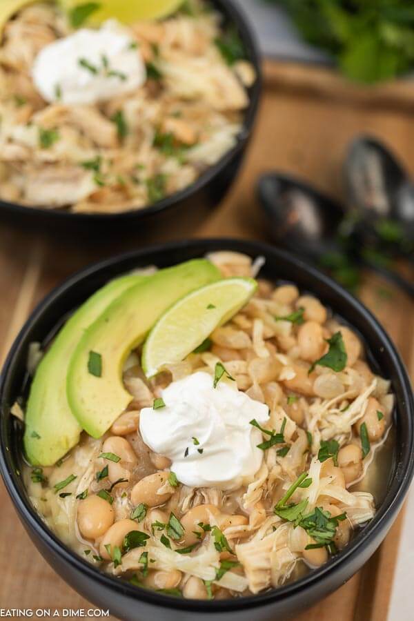 Close up image of two black bowls of white chicken chili with avocado and sour cream on a board. 